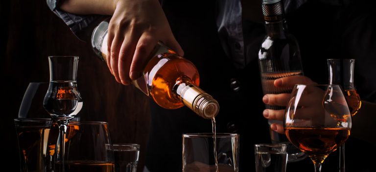 The bartender pours the dark golden rum in glass on the old bar counter. Vintage wooden background in pub or bar, night mood. Place for text, toning, selective focus